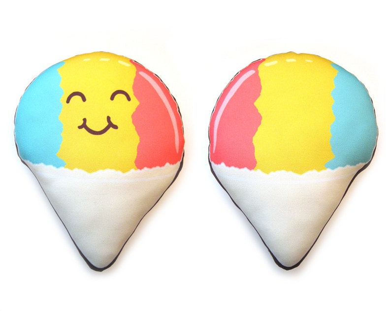 Puede incluir: Dos cojines de felpa con forma de cono de nieve. Uno tiene una cara sonriente, con secciones azules, amarillas y rojas sobre una base blanca. El otro coj&iacute;n tiene el mismo patr&oacute;n de color sin cara. Ambos tienen un contorno negro.