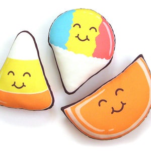 Puede incluir: Tres cojines de felpa con forma de comida: un caramelo de ma&iacute;z, un cono de nieve y una rodaja de naranja. Cada coj&iacute;n tiene una cara sonriente y est&aacute; delineado en marr&oacute;n. El caramelo de ma&iacute;z es amarillo, naranja y blanco. El cono de nieve es azul, amarillo, rojo y blanco. La rodaja de naranja es naranja.