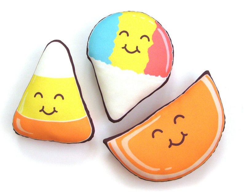 Puede incluir: Tres cojines de felpa con forma de comida: un caramelo de ma&iacute;z, un cono de nieve y una rodaja de naranja. Cada coj&iacute;n tiene una cara sonriente y est&aacute; delineado en marr&oacute;n. El caramelo de ma&iacute;z es amarillo, naranja y blanco. El cono de nieve es azul, amarillo, rojo y blanco. La rodaja de naranja es naranja.