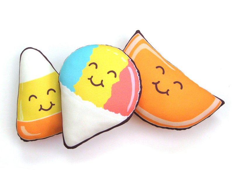 Puede incluir: Tres peluches de comida: un caramelo de ma&iacute;z, un cono de nieve y una rodaja de naranja. Cada uno tiene una cara sonriente. El caramelo de ma&iacute;z es amarillo, naranja y blanco. El cono de nieve es azul, amarillo y rosa. La rodaja de naranja es naranja.