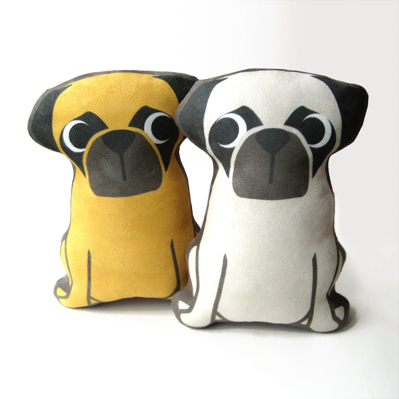 Puede incluir: Dos almohadas de peluche con forma de perro pug, una amarilla y otra blanca, con ojos y nariz negros. Las almohadas est&aacute;n sobre una superficie blanca.