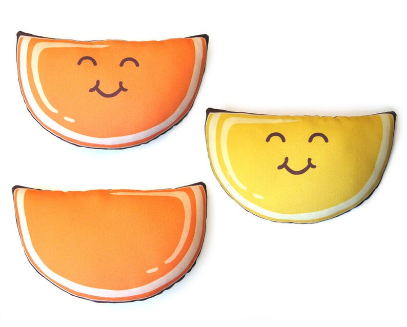 Puede incluir: Tres cojines de felpa con forma de rodajas de fruta. Dos rodajas de naranja y una rodaja de lim&oacute;n amarillo, cada una con una cara sonriente. Los cojines tienen un borde blanco y un contorno marr&oacute;n. Los cojines est&aacute;n sobre un fondo blanco.