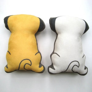 Puede incluir: Dos almohadas de peluche con forma de perro, una amarilla y otra blanca, con orejas y colas negras. Las almohadas est&aacute;n sobre una superficie blanca.