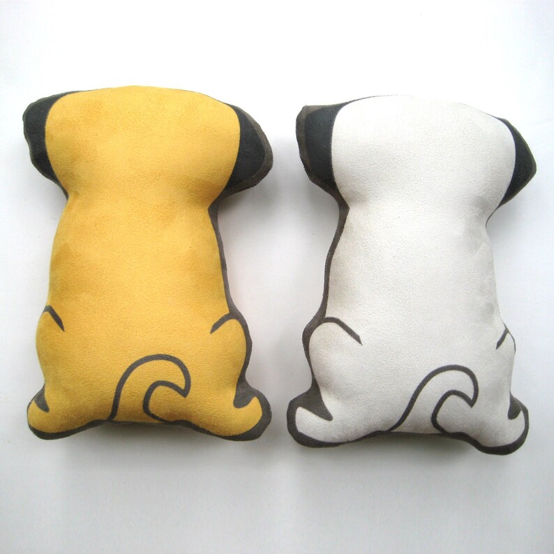 Puede incluir: Dos almohadas de peluche con forma de perro, una amarilla y otra blanca, con orejas y colas negras. Las almohadas est&aacute;n sobre una superficie blanca.