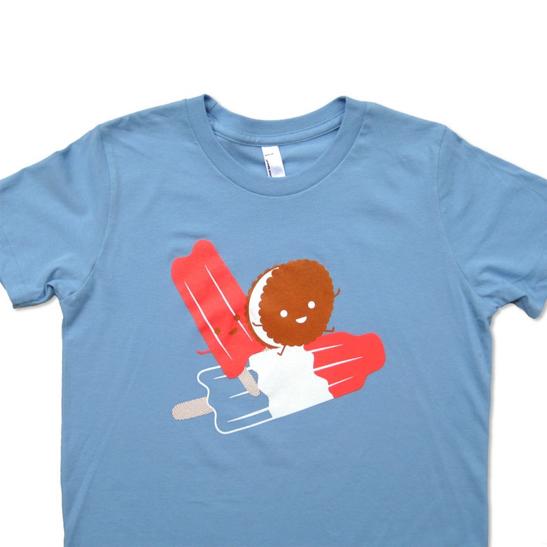May include: A light blue t-shirt with a graphic of a smiling chocolate cookie riding on a red, white, and blue popsicle.