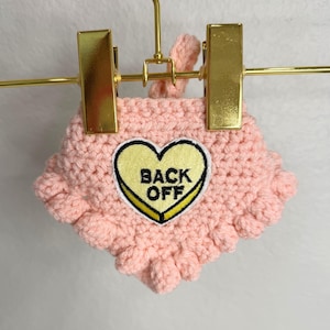 May include: A light pink crocheted garment featuring a textured bobble stitch pattern and a central yellow heart-shaped patch with a black border. The patch contains the text BACK OFF in bold black capital letters, centered on the heart.