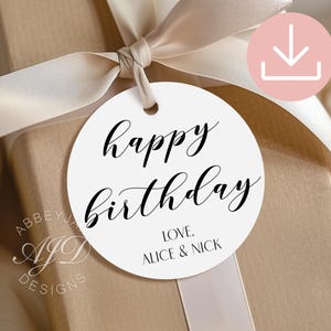 May include: A round white gift tag with black text that reads happy birthday and LOVE, ALICE & NICK. The tag is attached to a tan gift box with a light beige ribbon tied in a bow. A circular pink download icon is visible in the upper right corner.