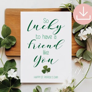 May include: A white card with green text that reads "So lucky to have a friend like you" and "Happy St. Patrick's Day!" A green shamrock is also featured. The card is on a wooden surface, surrounded by greenery and white flowers.