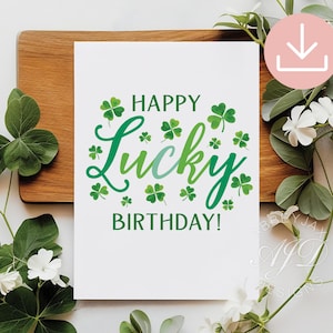 Puede incluir: Tarjeta de felicitación blanca con la frase "HAPPY LUCKY BIRTHDAY!" en verde, adornada con tréboles. La tarjeta está sobre una tabla de madera, con hojas verdes y flores blancas en el fondo.