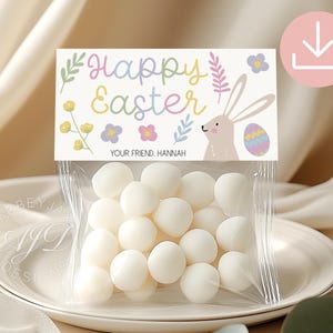 May include: A cellophane bag filled with white sweets, presented on a white plate. The bag is topped with a card featuring the words "Happy Easter" in pastel colours, a bunny, and an Easter egg illustration. The text "YOUR FRIEND, HANNAH" is also present.