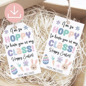 May include: Two white gift tags with the text "I'm so HOPPY to have you in my CLASS! Happy Easter!" in pastel colours. Each tag features Easter-themed illustrations of an egg, carrot, butterfly, and bunny. The tags are inside a cardboard box.