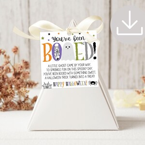 May include: A square white gift tag with a cream ribbon, featuring the text You've been BOOED! with a ghost, spider web, and spider graphic. The tag includes a poem about Halloween treats and the phrase Happy Halloween in colorful lettering.