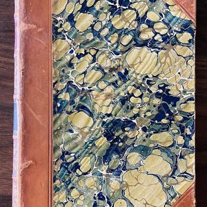 May include: A vintage book with a marbled cover in shades of blue, green, and gold. The book is bound in brown leather with a decorative spine.