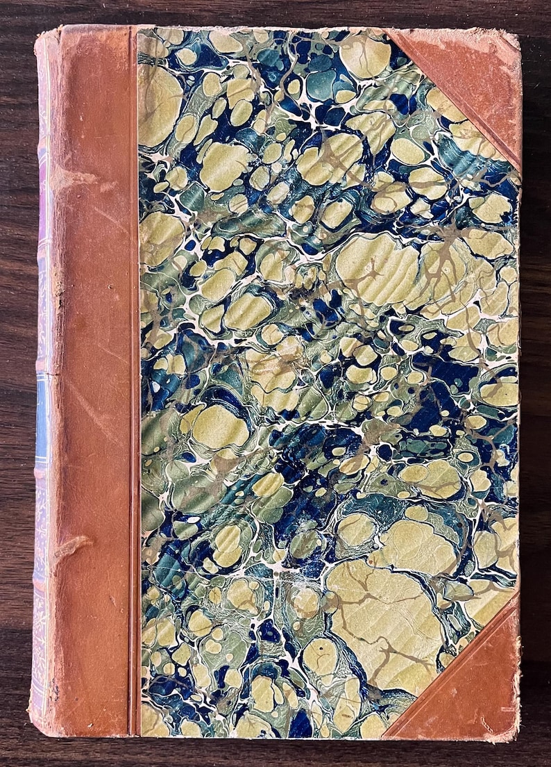 May include: A vintage book with a marbled cover in shades of blue, green, and gold. The book is bound in brown leather with a decorative spine.