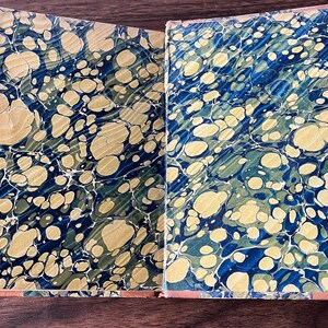 May include: A vintage book with a marbled cover in shades of blue, green, and gold. The book is open to reveal the marbled endpapers.