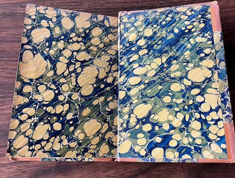 May include: A vintage book with a marbled cover in shades of blue, green, and gold. The book is open to reveal the marbled endpapers.