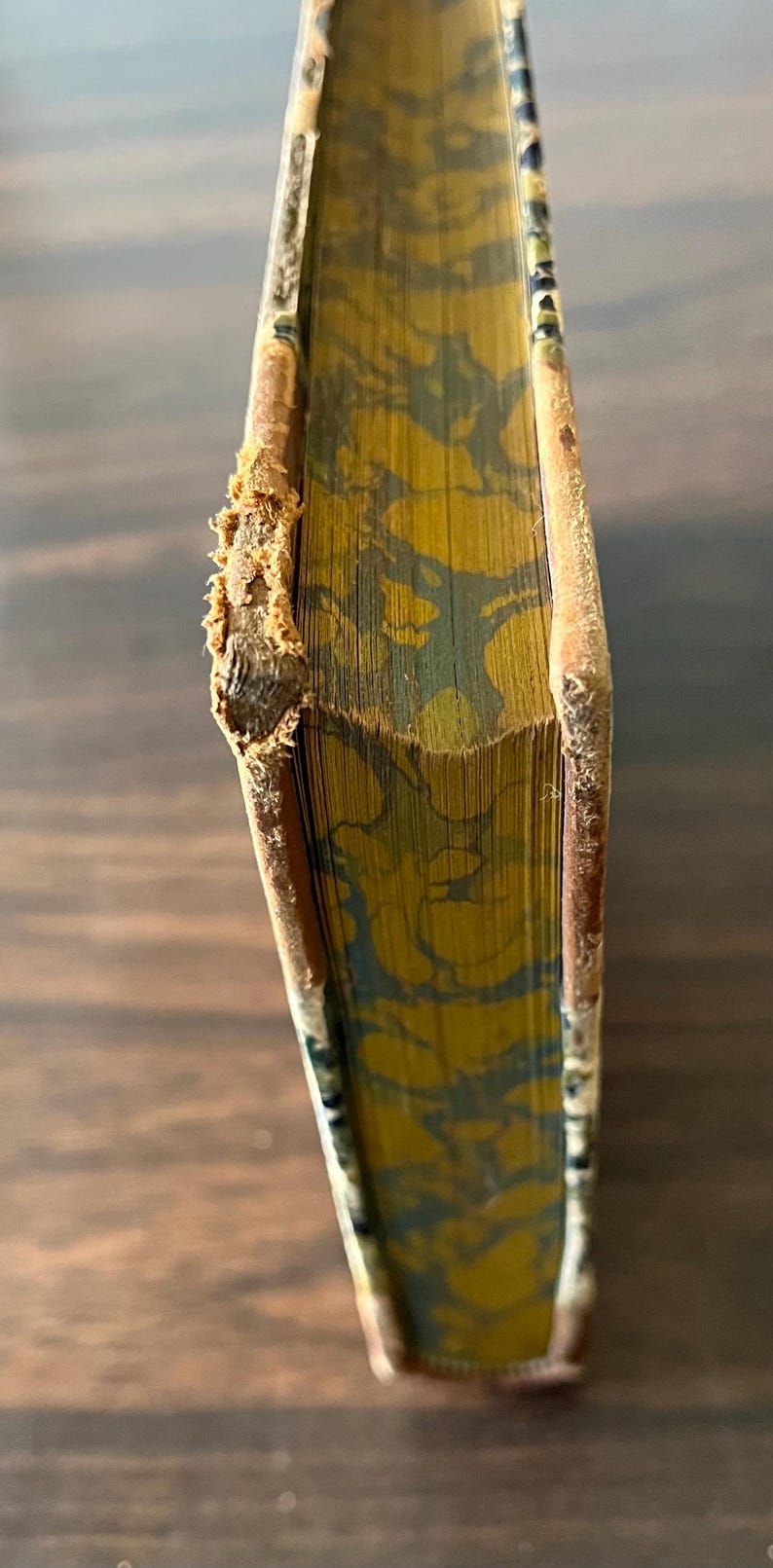 May include: Close-up of the spine of a vintage book with a marbled cover in shades of yellow and green. The book is bound in brown leather with a worn edge.