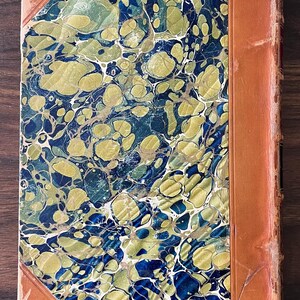 May include: A vintage book with a marbled cover in shades of blue, green, and gold. The book has a brown leather spine and is bound in a decorative style.