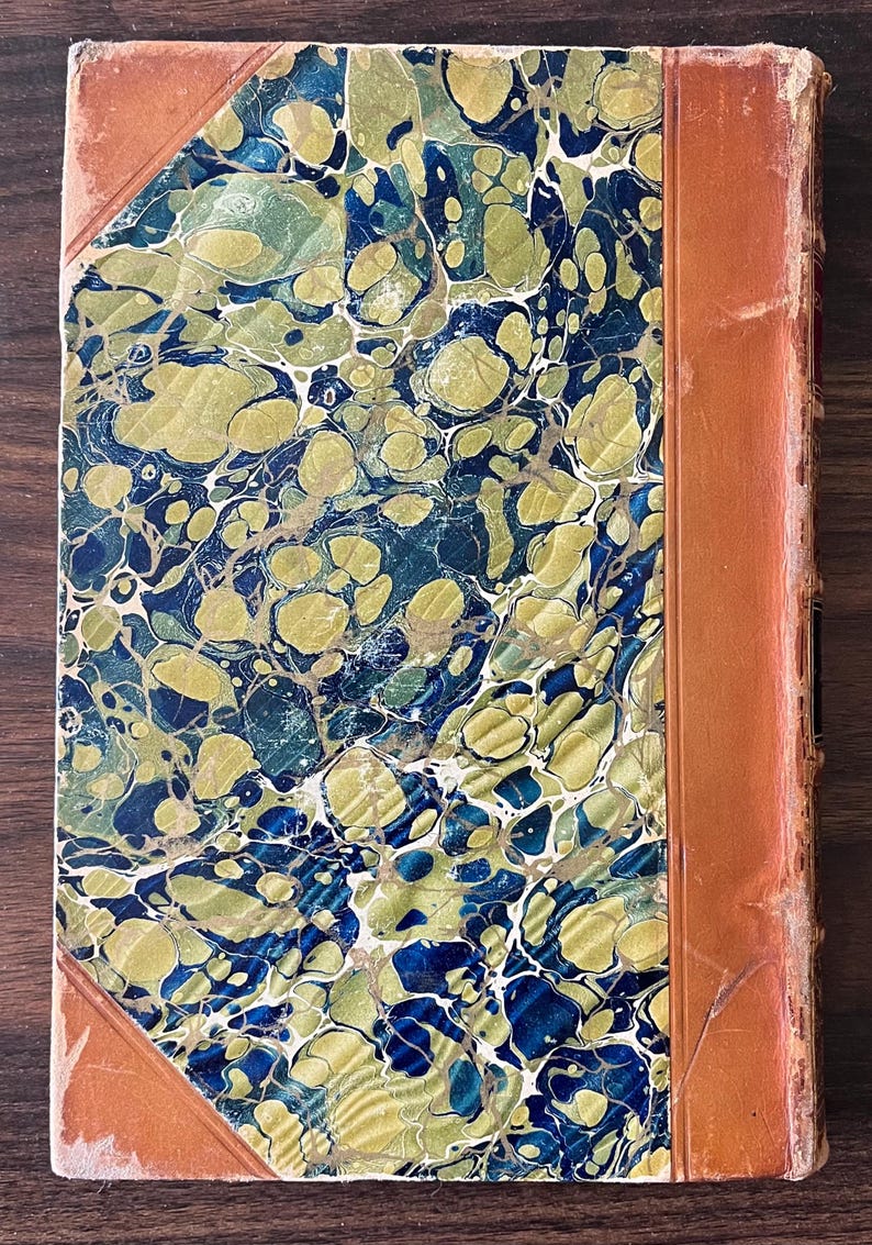 May include: A vintage book with a marbled cover in shades of blue, green, and gold. The book has a brown leather spine and is bound in a decorative style.