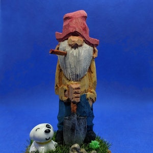 Op de afbeelding: Een houten beeldje van een kabouter met een rode muts en een schop in zijn hand. De kabouter heeft een witte baard en rookt een pijp. De kabouter staat op een houten voet met groen gras en een klein wit hondje.