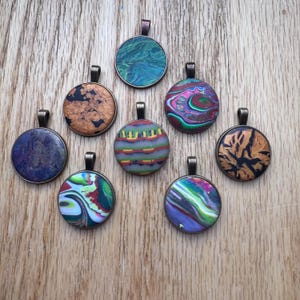 May include: A collection of eight round pendants with unique, colorful designs. Each pendant has a bronze-colored bail. The designs include swirls of blue, green, red, and purple, as well as textured patterns in brown and gold. The pendants are displayed on a wooden surface.