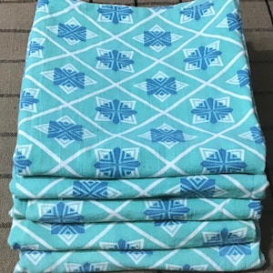 以下が含まれることがあります： ターコイズ色の折り畳まれた布が5枚重ねられています。各布には、白いひし形と青い花のようなデザインが繰り返されています。布はきちんと積み重ねられており、詳細なパターンを示しています。