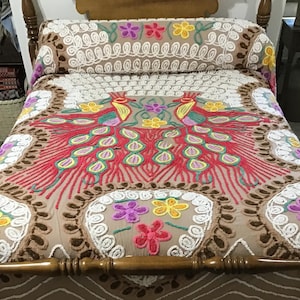 May include: A beige bedspread with a colorful embroidered peacock design. The bedspread features red, pink, green, yellow, and purple floral accents. The headboard cover has a repeating white swirl pattern with colorful flowers.