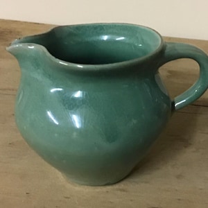 May include: A handcrafted, teal-colored ceramic pitcher with a rounded body, a curved handle, and a pouring spout. The pitcher has a glossy finish and sits on a wooden surface. This decorative pitcher is suitable for serving beverages.
