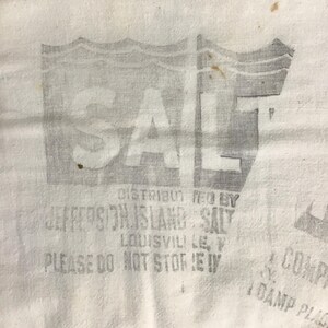May include: Close-up of a white textile with a faded gray print. The print features the word "SALT" in large capital letters, with additional text below indicating the distributor and location. The textile has a slightly aged appearance.