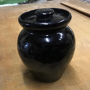 May include: A glossy black ceramic jar with a lid. The jar has a rounded body and a wide, slightly flared rim. The lid is a small, rounded knob. The jar is sitting on a light brown surface.