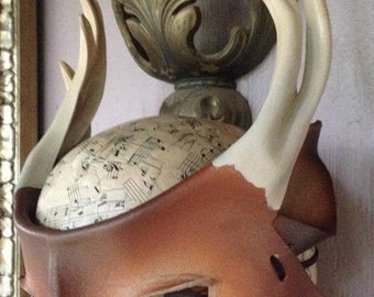 May include: A brown leather masquerade mask with a single eye hole and a white painted eye. The mask is decorated with two white antlers.