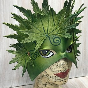Op de afbeelding: Een groen leren masker voor een maskerade met een bladontwerp. Het masker heeft groene ogen en een swirl-ontwerp op het voorhoofd. Het masker zit op een mannequinhoofd.