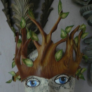 May include: A brown leather mask with a tree branch design and green leaves. The mask has two blue eyes and is worn on a mannequin head.