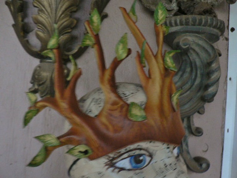 May include: A brown leather mask with a tree branch design and green leaves. The mask has a face with a painted eye and a textured background.