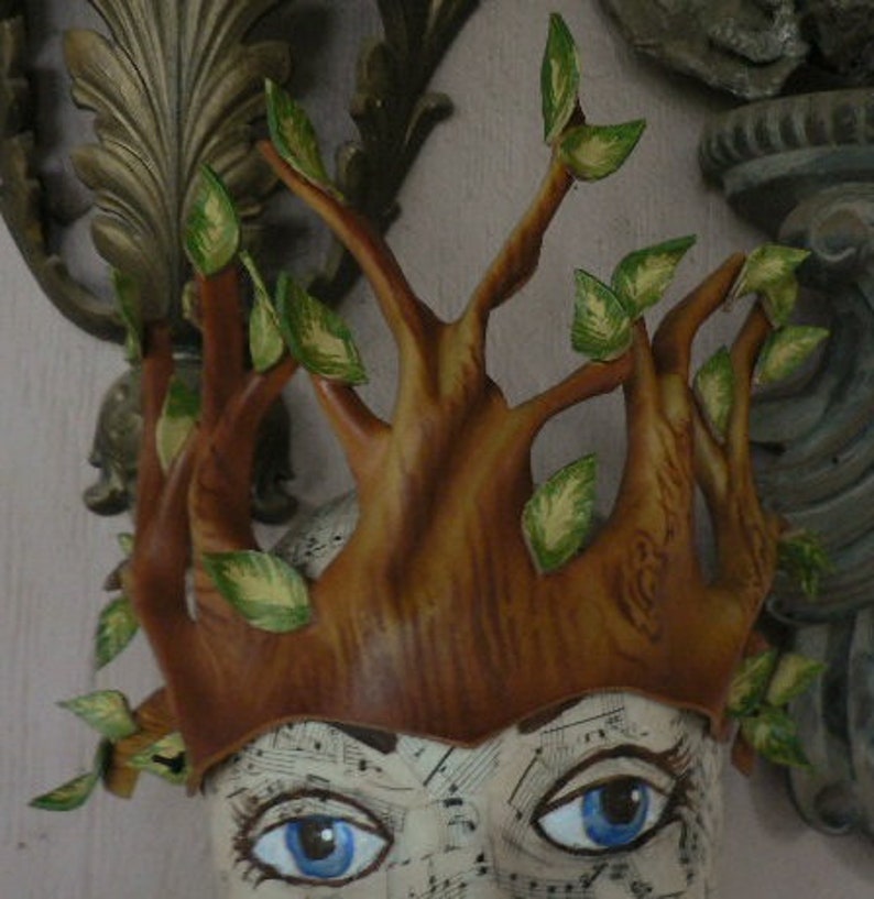 May include: A brown leather crown with green leaves, resembling a tree, worn on a mannequin head.