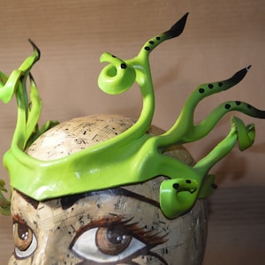 May include: A green and black headband with a unique, organic design. The headband is made of a flexible material and has a textured surface. It is decorated with black dots and has a pointed, curved shape.