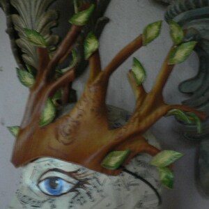 May include: A brown and green tree branch crown with leaves, made of paper mache, with a blue eye painted on a white background.