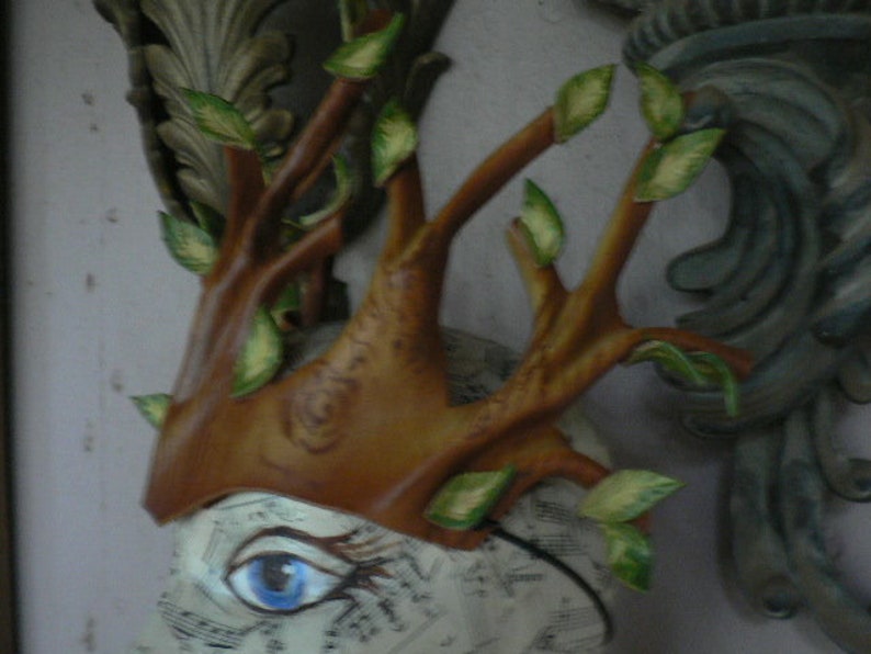 May include: A brown and green tree branch crown with leaves, made of paper mache, with a blue eye painted on a white background.