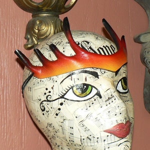 May include: A white mannequin head with a red and orange leather crown with points. The mannequin's face is decorated with black musical notes and a green eye.