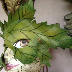 Puede incluir: Una máscara de cuero verde con forma de hoja con un ojo pintado y un acento marrón. La máscara está en una cabeza de maniquí con un fondo blanco.