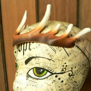 Peut inclure: Un serre-tête en bois de cerf blanc et brun avec une bande en cuir marron. Le serre-tête est porté sur une tête de mannequin recouverte de partitions musicales. Le mannequin a un œil peint avec du fard à paupières vert.