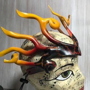 Könnte beinhalten: Ein handgefertigtes Kopfstück mit einem flammenartigen Design. Das Kopfstück weist gebogene, hornartige Elemente in Gelb-, Orange- und Rottönen auf, die an der Basis in Schwarz übergehen. Die Basis ist ein Band mit einem dekorativen Muster, und der Kopf ist mit Noten bedeckt.