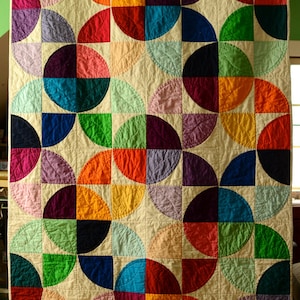 Könnte beinhalten: Eine handgefertigte Steppdecke mit einem lebendigen, geometrischen Design. Die Steppdecke zeigt ein sich wiederholendes Muster aus farbenfrohen Halbkreisen in Rot-, Orange-, Gelb-, Grün-, Blau-, Lila- und Rosatönen auf cremefarbenem Hintergrund. Die Steppdecke ist ein Patchwork-Design.