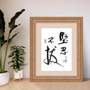 May include: Framed artwork featuring black calligraphy on a white background. The calligraphy includes several Chinese characters and a red stamp. The frame is gold with a detailed pattern. Two potted plants are visible in the background.