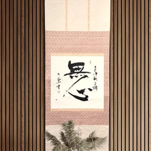 May include: A vertical scroll with Japanese calligraphy in black ink, mounted on a pink and cream background. The scroll hangs against a wooden slatted wall. A small vase with greenery sits on a wooden table below.