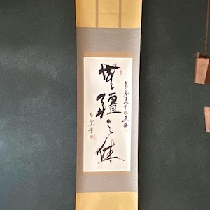 Könnte beinhalten: Eine traditionelle japanische Schriftrolle mit Kalligraphie, die an einer dunkelgrünen Wand hängt. Die Schriftrolle ist cremefarben mit goldenen Akzenten und einer Holzleiste am unteren Rand. Rechts hängt ein kleines hölzernes Flaschenzugsystem mit zwei Eimern.