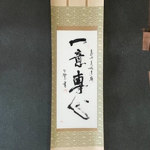 Puede incluir: Un rollo japonés tradicional con caligrafía negra, colgado contra una pared oscura. El rollo es de color crema con un fondo estampado y una barra de madera roja en la parte inferior. Un pequeño sistema de poleas de madera cuelga a la derecha.