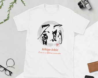 May include: A white short-sleeve t-shirt featuring a black Japanese calligraphy design inside a grey circular brushstroke. Below the graphic, the text reads Ichigo-Ichie and Once in a lifetime encounter in a red and black font.