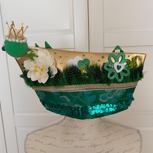 Könnte beinhalten: Dekorativer grüner und goldener Hut mit einem skurrilen Design. Zu den Merkmalen gehören eine goldene Krone, eine weiße Blume, grüne Federn und eine Verzierung zum Thema Frosch. Der Hut ist mit Pailletten und einer goldenen Bordüre verziert.