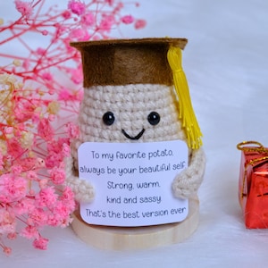 May include: A crocheted potato figure wearing a brown graduation cap with a yellow tassel. The potato holds a sign that reads, "To my favorite potato, always be your beautiful self. Strong, warm, kind and sassy. That's the best version ever."